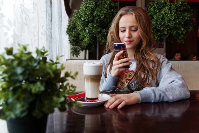 Dívka sedící u stolu s kávou a mobilem v ruce